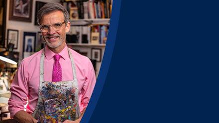 Dale Stephanos, a 2023 graduate from SNHU, standing in his studio and wearing an apron covered in paint.