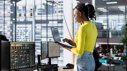 A woman working on a laptop surrounded by other computers and tech equipment to represent database administration.
