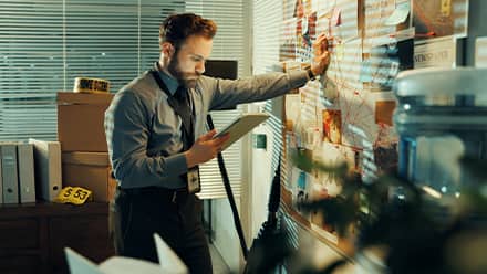 A detective with a criminal justice degree, leaning against a map and reviewing a case file in his hand.