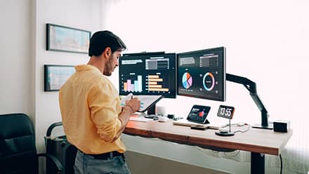A man in an orange shirt standing at a desk looking at two monitors and researching how to become an economist.