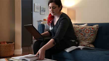 A master’s student sitting on the couch with a laptop and books, considering how long it will take her to finish her degree program.