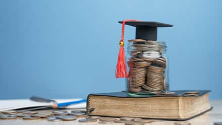 A book beneath a stack of coins with a tiny graduate cap on top.