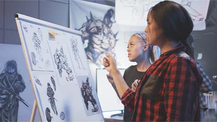 A game designer working with an artist to create a video game character concept using sketches and digital artwork in a design studio.