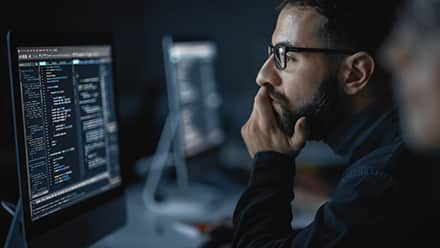 A man wearing glasses looks at code on a computer screen with his chin resting on his hand.