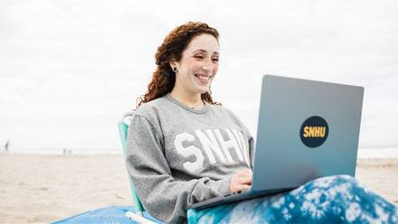 Mariel Embry, a 2022 SNHU graduate sitting on a beach with her laptop working on her liberal arts degree.