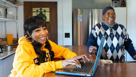 Remy Twine, a 2025 SNHU marketing graduate wearing a yellow SNHU sweatshirt working on a laptop at a wooden table inside a home, with another person seated nearby.