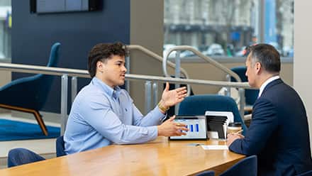Two people seated at a table during a business meeting, having a discussion, with a tablet displaying charts on the table between them.