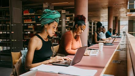 Two master's students studying in a library, one using a laptop and the other reading a textbook.
