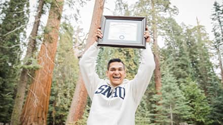 Juan Munoz, a 2022 and 2024 SNHU graduate, holding the diploma for his MBA over his head with pine trees behind him.