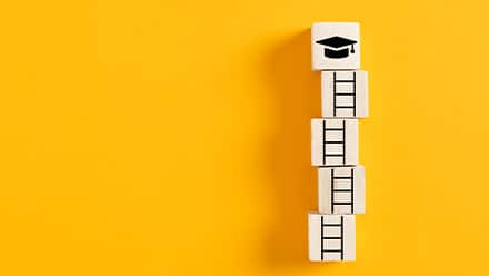 A stack of five building blocks on a yellow backdrop: the bottom four blocks feature pieces of a ladder and the top depicts a graduation cap