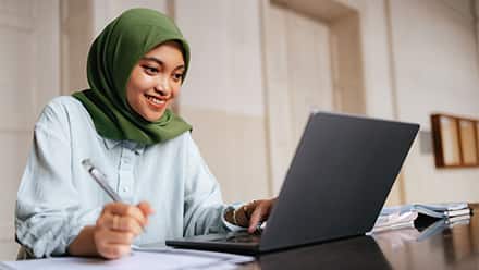 A smiling woman wearing a green hijab and a button-down shirt sitting at a laptop with a pen in hand.