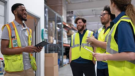 A supply chain manager holding a tablet, speaking with three other professionals wearing fluorescent yellow vests and working in a warehouse.