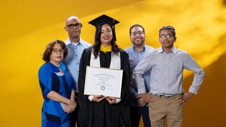 Brendaliz De Los Santos, center, dressed in a graduation cap and gown and holding her SNHU diploma, surrounded by 4 family members who supported her in college.