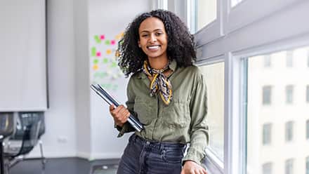 A woman smiling while holding a laptop and notebooks in a modern office.