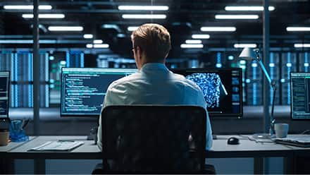 The back of a professional with a master's in cybersecurity at work, sitting at a desk in a large office building with two monitors in front of him.
