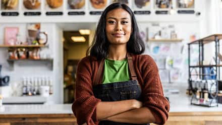 An entrepreneur wearing an apron standing in front of a desk in her business.