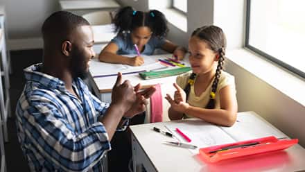 A male teacher kneeling and making eye contact while instructing a child in a classroom.