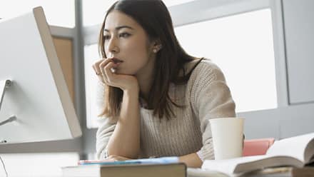 A college student searching for different types of scholarships on a desktop computer with a coffee cup and notebook open beside her.