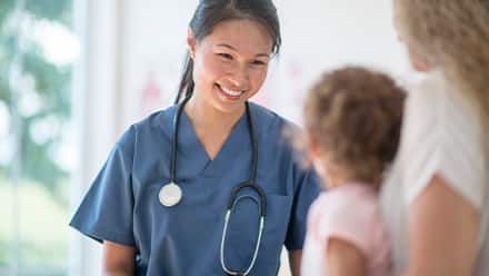 A nursing professional that finished the degree needed to become a nurse, wearing blue scrubs and a stethoscope and meeting with a pediatric patient.