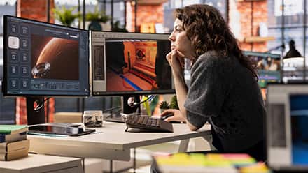 A game designer sitting in front of two monitors with a video game development software open, showing a planet and the inside of a spacecraft.
