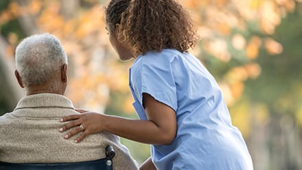 A CNA wearing blue scrubs with her hand on the shoulder of a patient in a wheelchair.