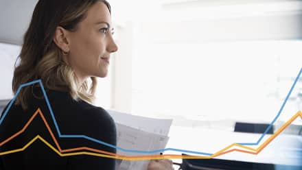 An economist reviewing paperwork at her desk with a blue, orange and yellow line graph overlay.