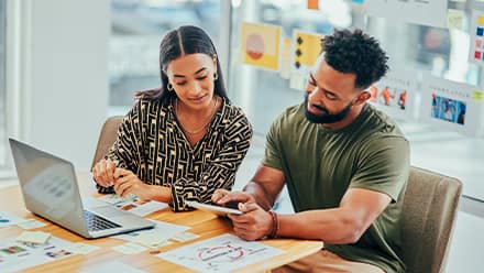 Two brand managers sitting together and looking at a tablet with printed documents and a laptop open in front of them.