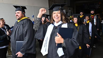 A graduate wearing a first generation stoll raising a fist and smiling while walking in a line of other graduates holding their degrees.