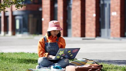 A Gen Z college student sitting in a grassy area while doing schoolwork on her laptop.