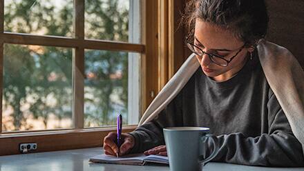 A speculative fiction writer with a mug, writing in a notebook beside a window overlooking a lake.