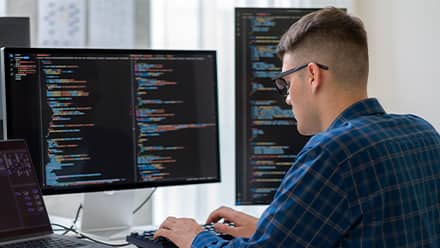 A computer science student with a laptop and two monitors of code in front of him, considering what master's degree to get.