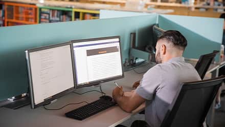 A transfer student sits in a library working on online coursework using two computer monitors.