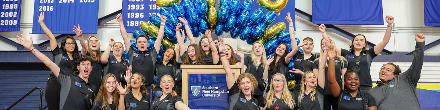 SNHU students celebrating around a podium. Behind them is an arch of balloons in SNHU colors.