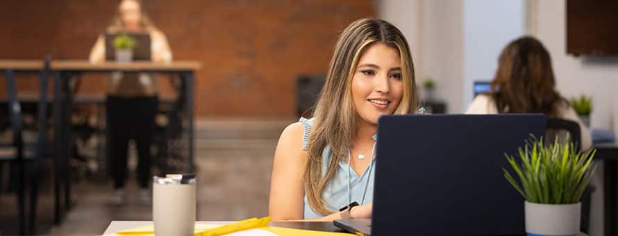 An SNHU student working on her laptop in a community space and a mug of coffee next to her.