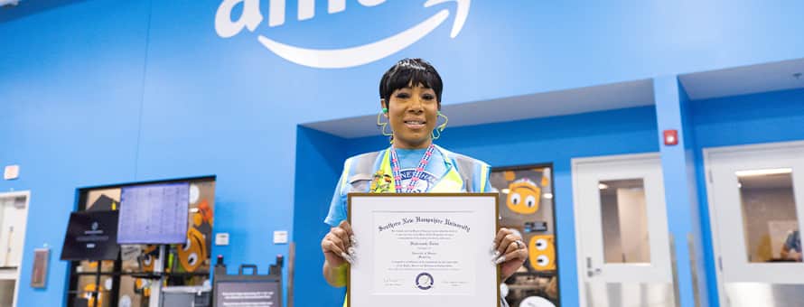 SNHU graduate, Remy Twine, standing in an Amazon facility holding her SNHU diploma.