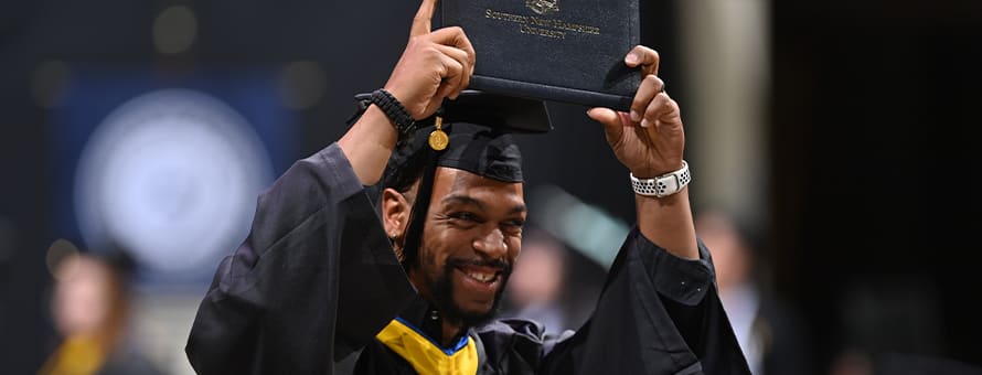 A graduate dressed in a cap and gown and holding a diploma over his head at the Fall 2023 SNHU Commencement.