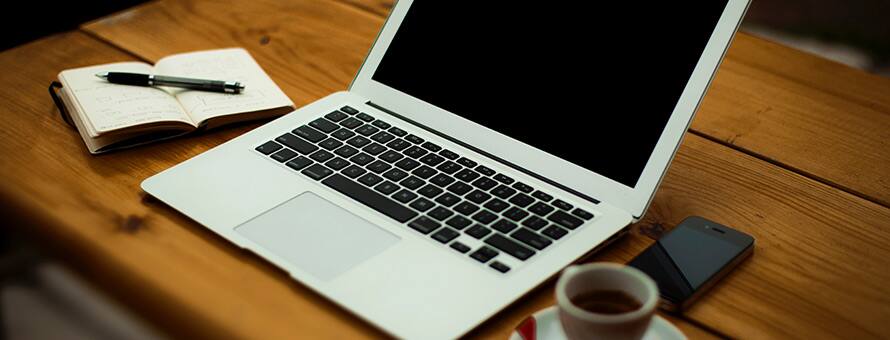 Pencil resting on a small open notebook next to a laptop and cup of coffee on a table