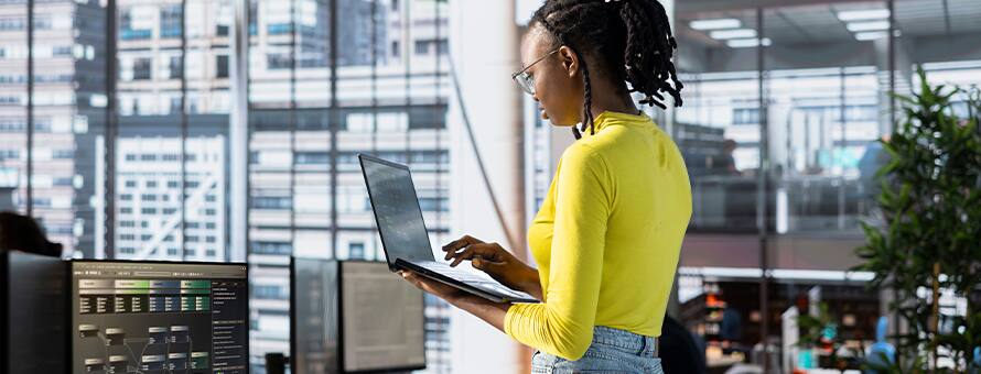 A woman working on a laptop surrounded by other computers and tech equipment to represent database administration.