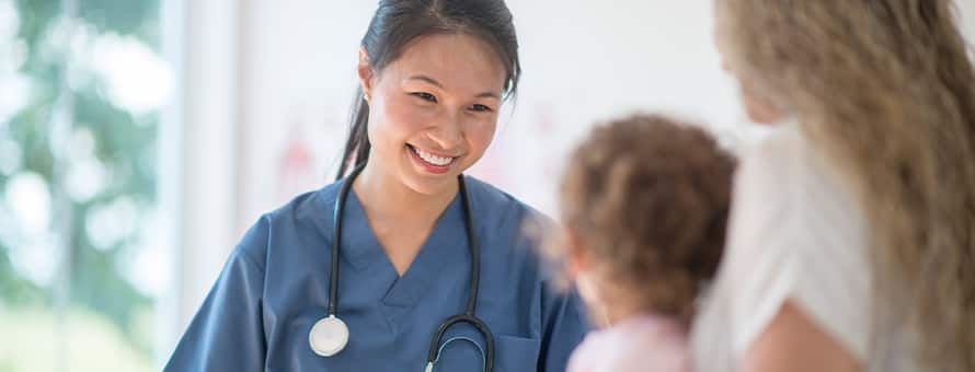 A nursing professional that finished the degree needed to become a nurse, wearing blue scrubs and a stethoscope and meeting with a pediatric patient.