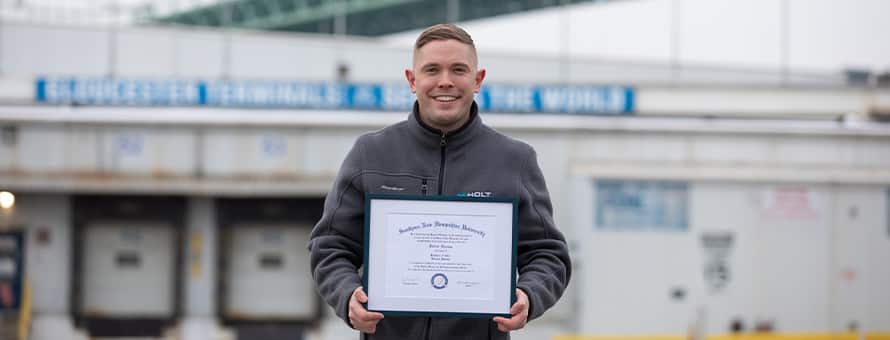 Rob Thomas, a 2024 SNHU graduate holding his diploma standing outside in an industrial area.