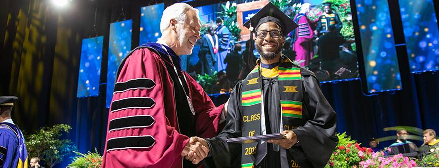 Man in a graduation gown shaking hands with a university president as he gets his degree.