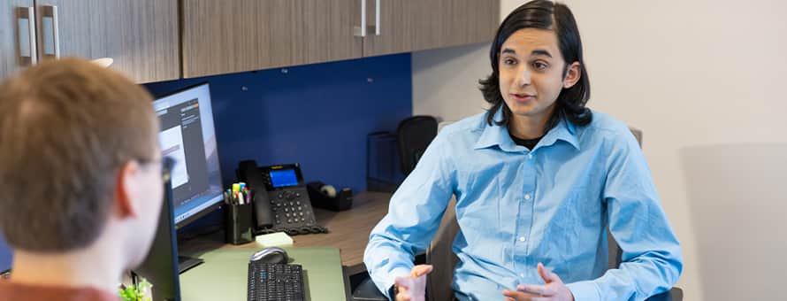 A hiring manager turned away from a computer, speaking to a job candidate about a technical interview.