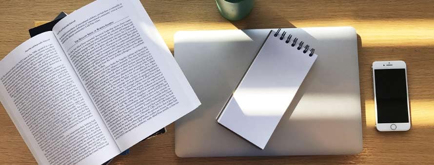 A textbook, notepad and laptop, travel mug and phone sitting on a desk for taking notes