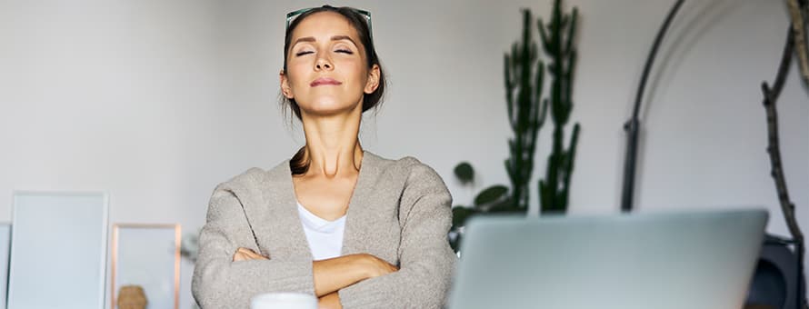 A woman sitting at her desk with her arms crossed and eyes closed practicing mindfulness meditation