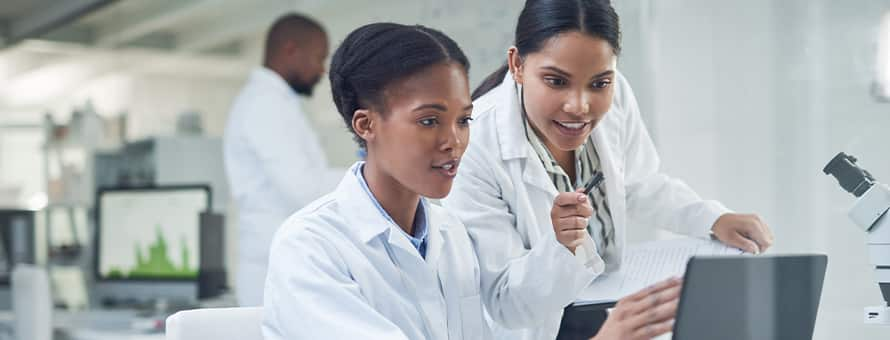 Two women in lab coats working in health science looking at a computer with a co-worker in the lab behind them