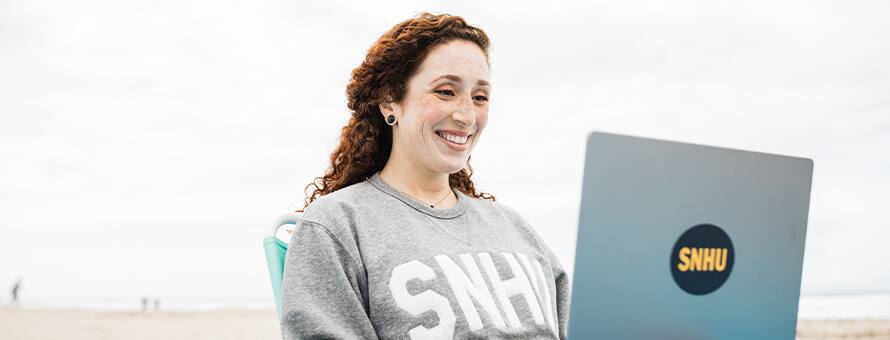 Mariel Embry, a 2022 SNHU graduate sitting on a beach with her laptop working on her liberal arts degree.