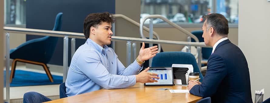 Two people seated at a table during a business meeting, having a discussion, with a tablet displaying charts on the table between them.
