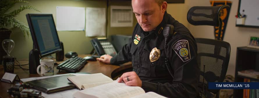 Tim McMillan in his police uniform working at his desk with text Tim McMillan '15