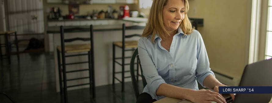 Woman sitting in her kitchen typing on a laptop, with text "Lori Sharp '14"