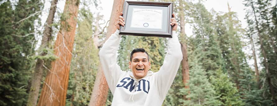 Juan Munoz, a 2022 and 2024 SNHU graduate, holding the diploma for his MBA over his head with pine trees behind him.